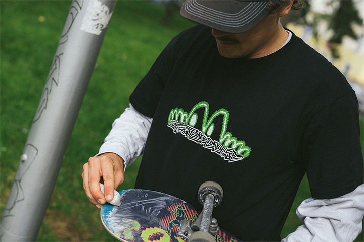 Person beim Wachsen eines Skateboards im Park, getragen wird ein schwarzes skatedeluxe T-Shirt mit neon-grünem Logo über einem weißen Longsleeve.