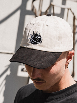 Crèmekleurige pet met zwarte klep en geborduurd patchdesign van skatedeluxe, gedragen in een stedelijke buitenomgeving.