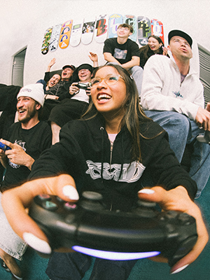 Een groep jongeren in skatedeluxe streetwear hoodies met opvallende prints zit op een bank. Op de voorgrond houdt iemand een gamecontroller vast. Op de achtergrond zijn skateboards te zien als muurdecoratie.