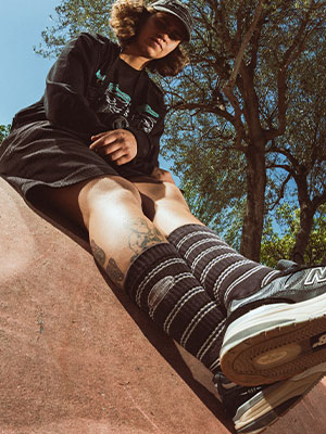 Low-angle lifestyle photo at the skate spot: striped skatedeluxe socks (black/grey) and skate shoes prominent, summer sunlight and natural scenery.