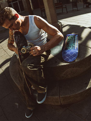 Pol Catena sits in the sunlight at a skate spot and unscrews a skateboard; to his right is a new skatedeluxe deck with blue graphics.