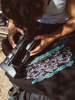 Close-up of camera and outfit: camcorder in the hands, a black skatedeluxe T-shirt with a large print of curved lines lying on the leg, outside in the sunlight.