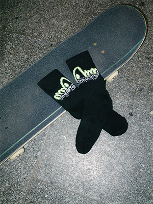 Black SKATEDELUXE socks with neon graffiti-style logo placed on a skateboard deck, photographed on a stone surface.