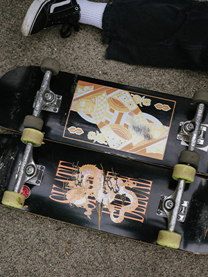 Two black SKATEDELUXE skateboards on pavement, one with a gold dragon graphic, the other with a geometric gold and white design, person with skate shoes nearby.