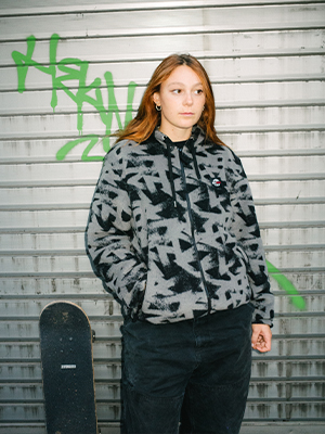 Person in grey and black SKATEDELUXE fleece hoodie with all-over graffiti print, black pants, standing by a metallic wall with green graffiti and skateboard.