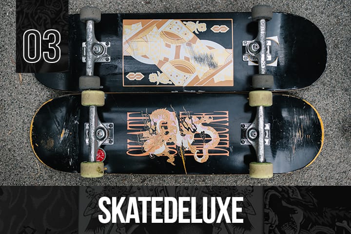 Two Skatedeluxe black skateboard decks with gold dragon and abstract graphics, fully assembled with trucks and wheels, displayed on asphalt.