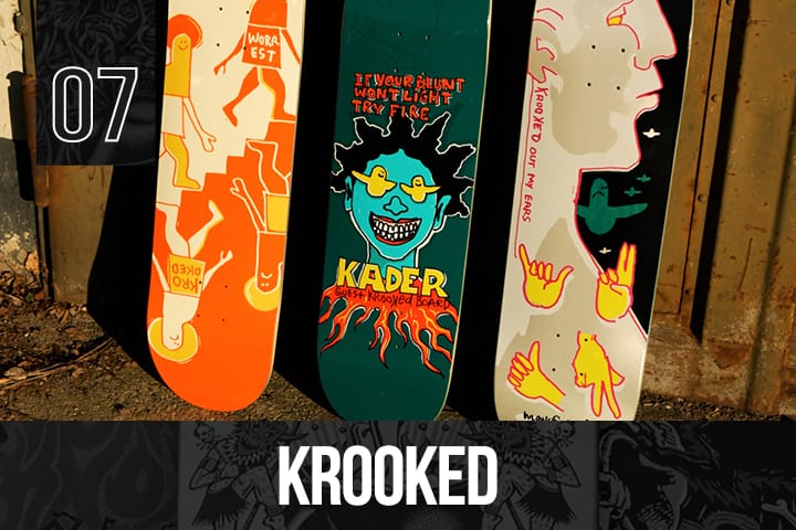 Three Krooked Skateboards decks with hand-drawn graphics: orange with figures, green with a blue face and flames, cream with yellow hands and a profile, arranged vertically.