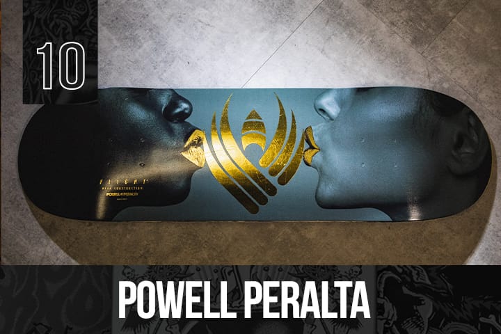 Powell Peralta skateboard deck with a sleek black base and metallic gold lips and emblem graphic, displayed on a grey floor.