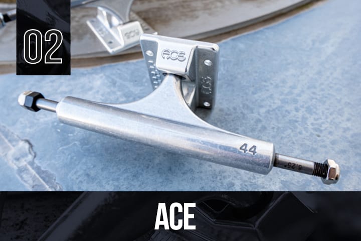 Silver Ace skateboard truck with "ACE" and "44" markings, placed on a blue metallic surface, another truck blurred in the background.