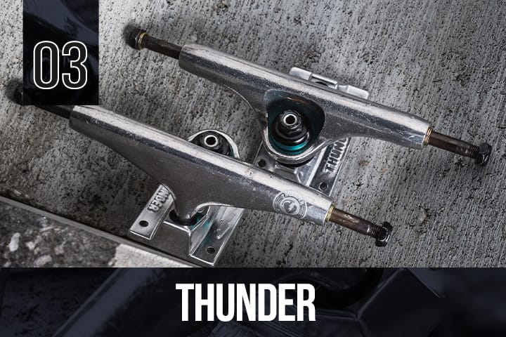 Two Thunder skateboard trucks in silver, positioned side by side on a rough grey concrete surface, showing visible branding and wear.