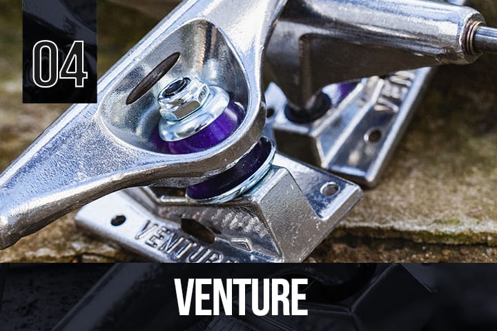 Close-up of Venture skateboard trucks in silver with purple bushings, "VENTURE" branding on the baseplate, placed on a rough stone surface.