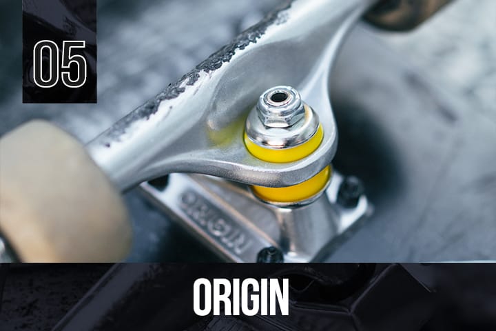 Close-up of Origin skateboard truck with bright yellow bushings, mounted on a skateboard, "ORIGIN" branding visible, blurred background.