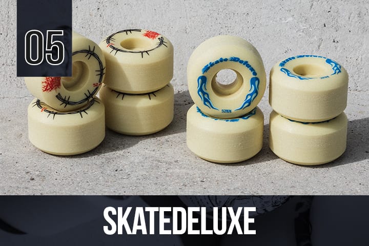 Skatedeluxe skateboard wheels in pale yellow with black barbed wire and blue flame graphics, stacked on a rough concrete surface.