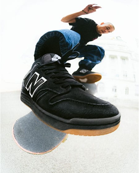 Dynamisches Skateboard-Foto: Ein schwarzer New Balance Numeric Skateschuh mit brauner Gummisohle füllt den unteren Bildbereich, darüber ist ein Skater mitten im Flip Trick in der Luft zu sehen, unscharfer Hintergrund mit heller Architektur.