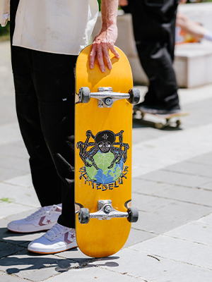 Geel skateboard met een schedel- en wereldbolontwerp van skatedeluxe, vastgehouden door een persoon in een zwarte broek en witte sneakers; stedelijke achtergrond met andere skateboarders.