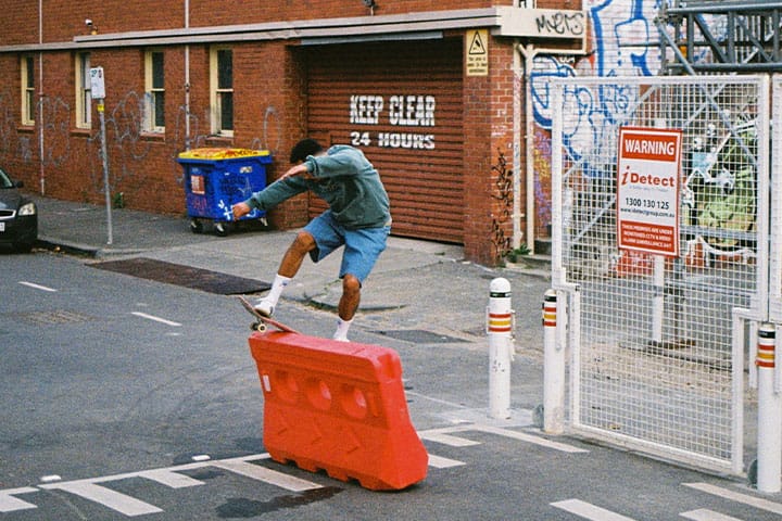 Person with denim shorts performs a skateboard trick over a red barrier, surrounded by an urban street environment.