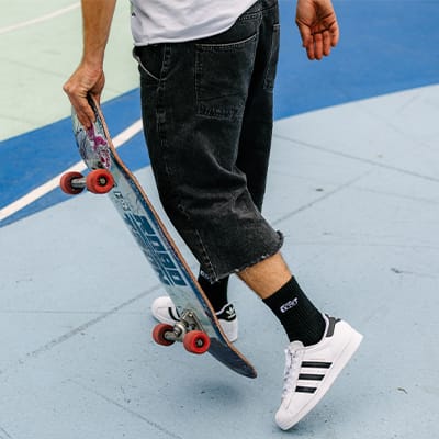 Skatewear scene with white sneakers, black denim shorts, and a skateboard in hand; the blue skatepark floor is visible in the background.