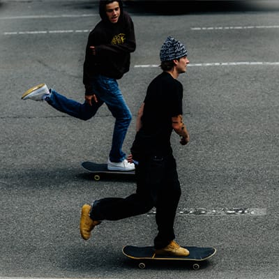 Urban streetwear motif with two skateboarders in motion, wearing black outfits and contrasting-colour shoes on a street.