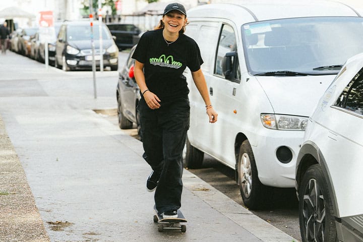 Female skateboarder in black skatedeluxe shirt and cap cruising down a city sidewalk in Vienna, surrounded by parked vehicles.