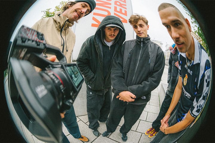 Skatedeluxe team in black streetwear posing for a fisheye camera in Vienna cityscape.