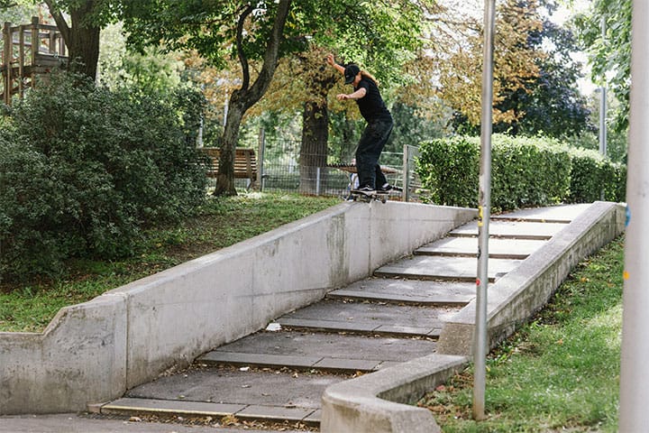 Skatedeluxe athlete skating a ledge with a 50/50 grind in a green urban park in Vienna, surrounded by trees and bushes.