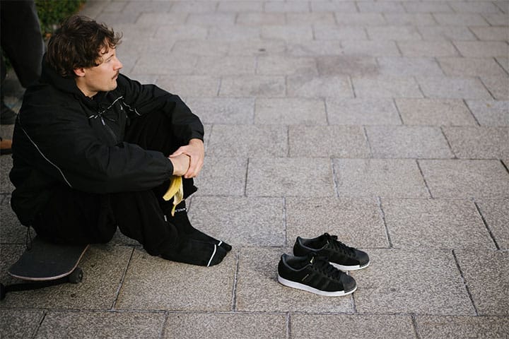 Person in black streetwear taking a break on a skateboard, holding a banana, shoes placed nearby on stone tiles.