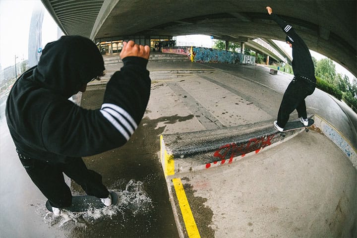 A skatedeluxe rider performing a ride on fs crooked on a wet spot under an urban overpass in Vienna, wearing black streetwear, landing in a puddle.