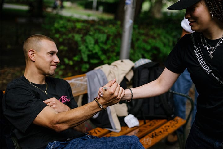 Two people shake hands on a park bench in Vienna, one wearing a skatedeluxe t-shirt, with backpacks in the background.