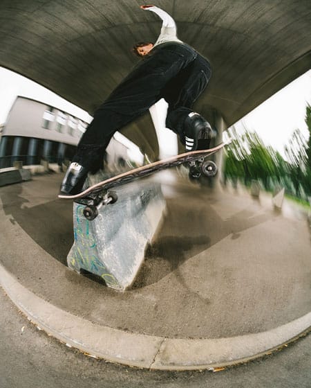 Skatedeluxe skateboarder in black streetwear skating a wet concrete ledge with yellow lines with a frontside noseslide pop out under a Vienna bridge.