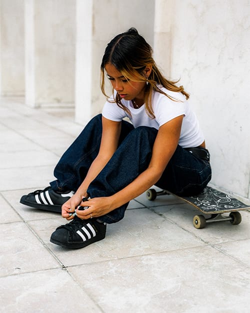 Nahaufnahme einer Skaterin, die in einem hellen Beton-Umfeld ihre schwarzen adidas Superstar ADV Skateschuhe mit Shell-Toe und wei&szlig;en Streifen zubindet, Skateboard im Hintergrund, Fokus auf On-Feet-Passform.