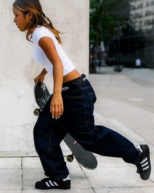 Seitliche Actionaufnahme einer Skaterin, die in schwarzen adidas Superstar ADV Skateschuhen mit wei&szlig;en Streifen auf einem Skateboard anst&ouml;&szlig;t und in einem urbanen Umfeld losf&auml;hrt.