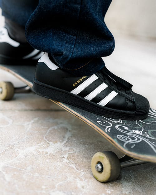 Close-up des adidas Superstar ADV Skateschuhs in Schwarz, platziert auf einem verzierten Skateboard-Deck auf hellem Steinboden, ideal zur Darstellung von Material, Passform und Boardkontrolle.