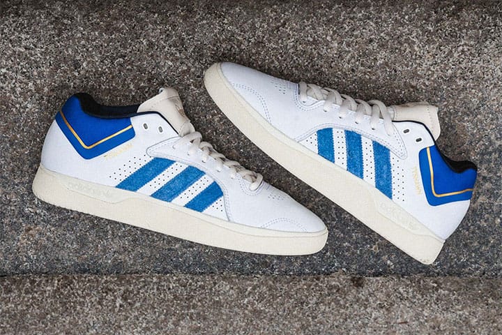 adidas Tyshawn signature sneakers in white with blue stripes and gold accents, placed on a concrete step.