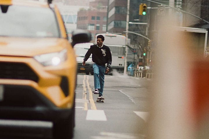 Tyshawn Jones patinando en una calle de la ciudad de Nueva York, vestido con una hoodie adidas negra con el logotipo blanco, vaqueros azul claro y zapatillas adidas Tyshawn II blancas, con un taxi amarillo en primer plano.