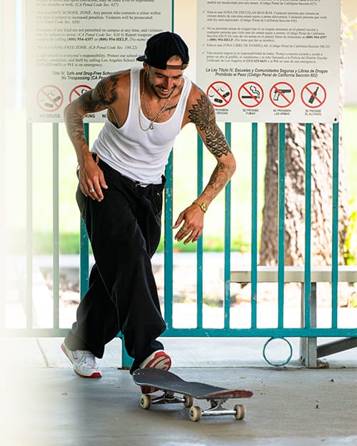 TJ Rogers wearing éS TJ Rogers white and red skate shoes, standing with skateboard in hand, urban background with signage, showcasing streetwear style and tattooed arms.
