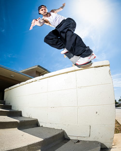 TJ Rogers performing a backside noseblunt on a white ledge, wearing éS TJ Rogers white and red skate shoes, sunny day, dynamic street skateboarding scene.