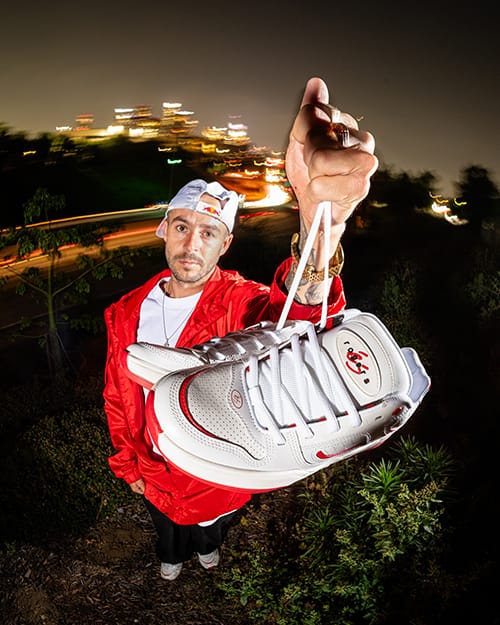 Close-up of TJ Rogers holding a pair of éS TJ Rogers white and red skate shoes towards the camera, detailed view of logo and design, blurred city lights in background.