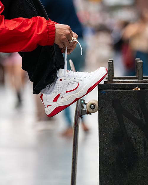 Detail shot of éS TJ Rogers pro model shoes, white with red accents, being tied by TJ Rogers in red jacket, city scene with blurred pedestrians.