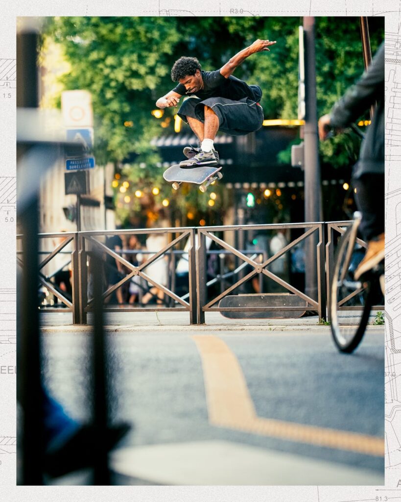 Skateboarder performing a flip trick over a metal street barrier in an urban setting, wearing black shorts, white socks, and New Balance Numeric 770 sneakers.