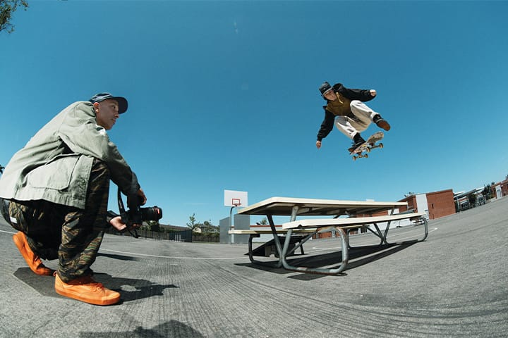 Skater Shiloh Catori performs a kickflip over a picnic table on an outdoor basketball court, photographed by Atiba Jefferson for Vans; bright blue sky, urban streetwear style, and orange skate shoes visible