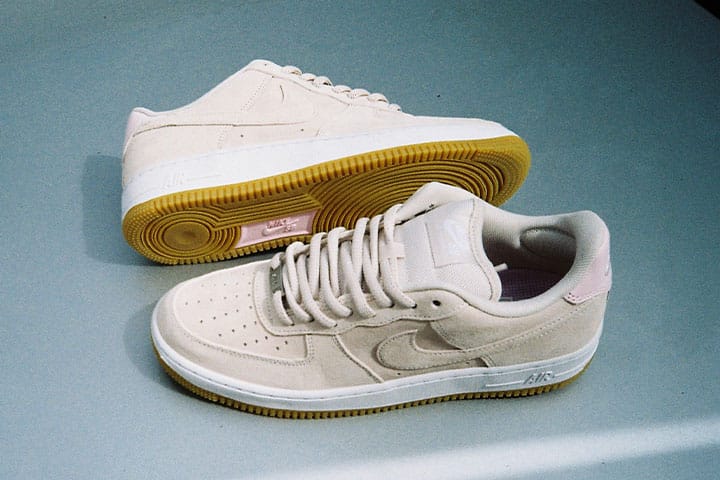 Zwei beigefarbene Nike SB Air Force 1 aus Wildleder, einer seitlich und einer mit sichtbarer Gummiaußensohle, auf glatter Oberfläche.