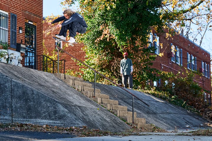 Ville Vester réalise un gros fakie ollie avec son skateboard au-dessus d’un handrail sur une rampe en béton raide, devant des maisons en briques rouges, tandis qu’une autre personne regarde depuis le bas du spot ; le trick montre la Nike SB AF-1 en action sur un spot de street classique.