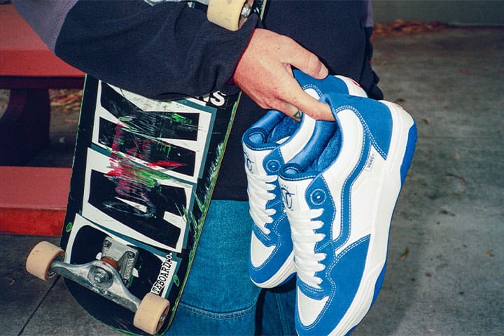 Un skater sostiene en la mano derecha un par de zapatillas de skate Vans Rowan 2 azules y blancas, junto a una tabla de skate Baker muy usada.