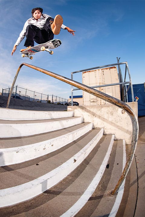 Dynamisches Skate-Foto, das Nick Michel in weiter Hose und Hemd zeigt, wie er in braunen Vans Z3 mit einem One Foot &uuml;ber eine Treppe und ein gebogenes Gel&auml;nder fliegt.