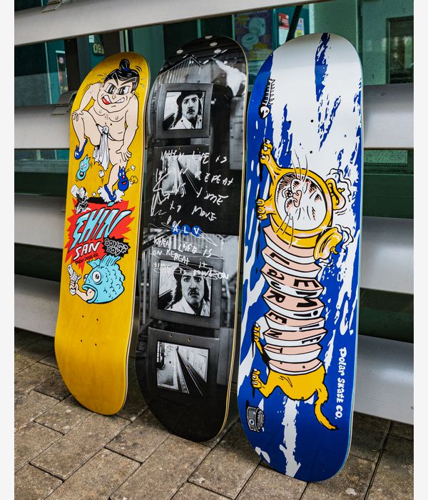 Photo illustrant le style de vie de la planche de skateboard « Polar Laurent Accordion Dog » de 8,375" (planche de skateboard) dans une rangée de trois ; planche de droite, de couleur bleue dominante, arborant un motif de chien de dessin animé au corps en accordéon jaune et blanc ; à côté, une planche jaune de dessin animé portant l'inscription rouge « Shiv Saw » ; et au centre, une planche présentant un collage photographique en noir et blanc, le tout sur fond de trottoir urbain et de garde-corps métallique.