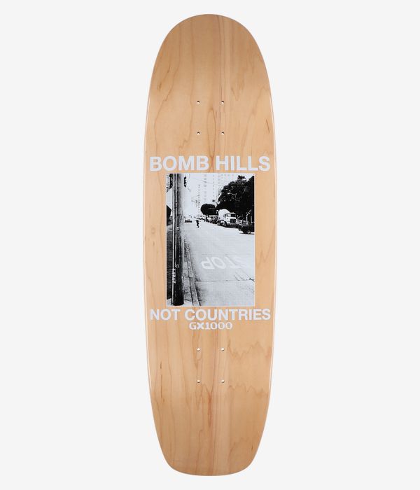 Planche de skateboard GX1000 Bomb Hills Cruiser 9 pouces, vue de dessous complète ; planche beige à motif bois avec, au centre, une photo en noir et blanc représentant une scène urbaine, ainsi que les inscriptions « BOMB HILLS » et « NOT COUNTRIES » en blanc ; image nette prise en studio ; fond blanc