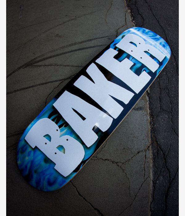Lifestyle image of Baker Zach Field Of Depth 8.25" skateboard deck with blue flame gradient edges, central black panel and large white BAKER block letters, photographed on cracked concrete/asphalt.