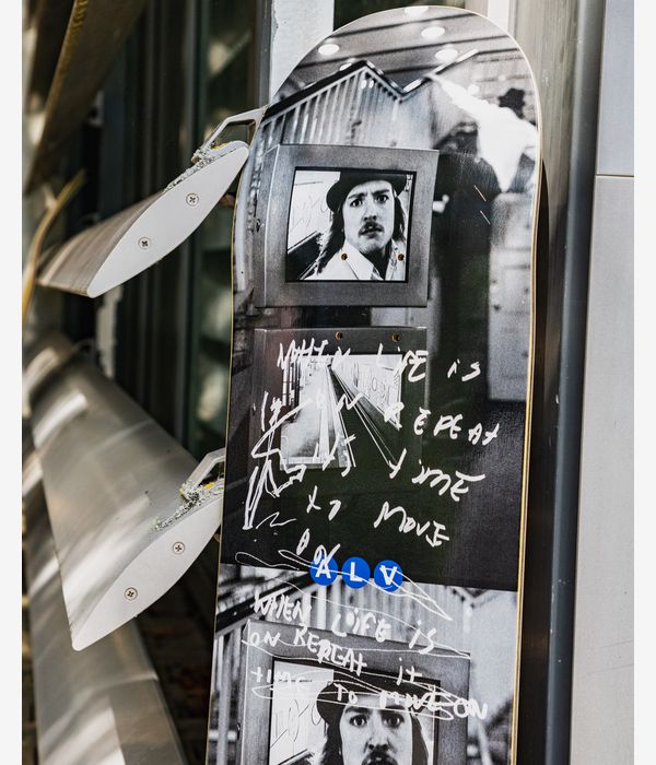 Polar Alv Märkisches Museum 8.75" skateboard deck — black top graphic with black-and-white photographic portraits, bold white handwritten script and a small cluster of blue stickers; leaned against metal fixtures, lifestyle image